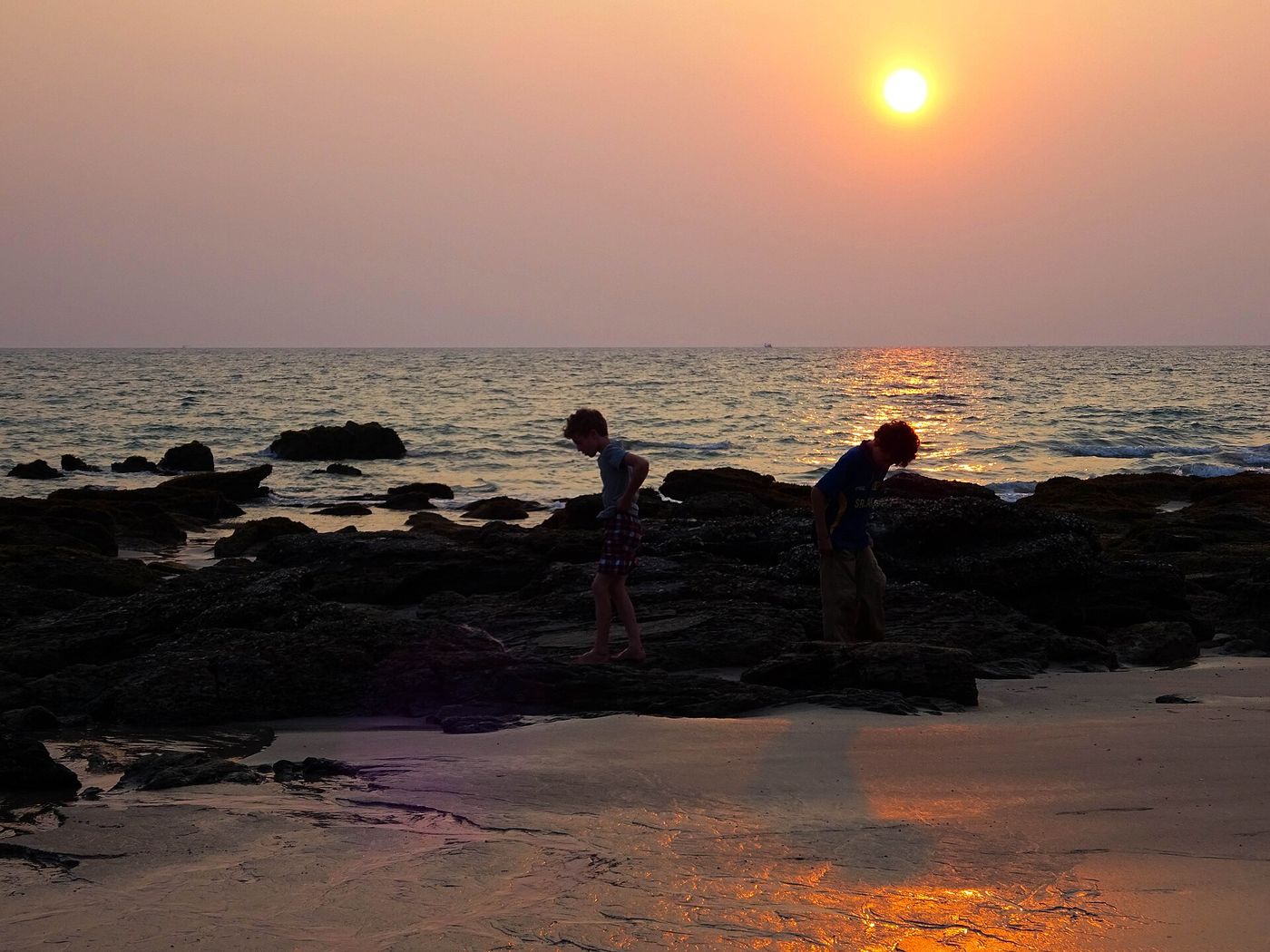 Two boys hunting for crabs among the dark rocks at low tide as the sun sets over a pink sky on Koh Lanta